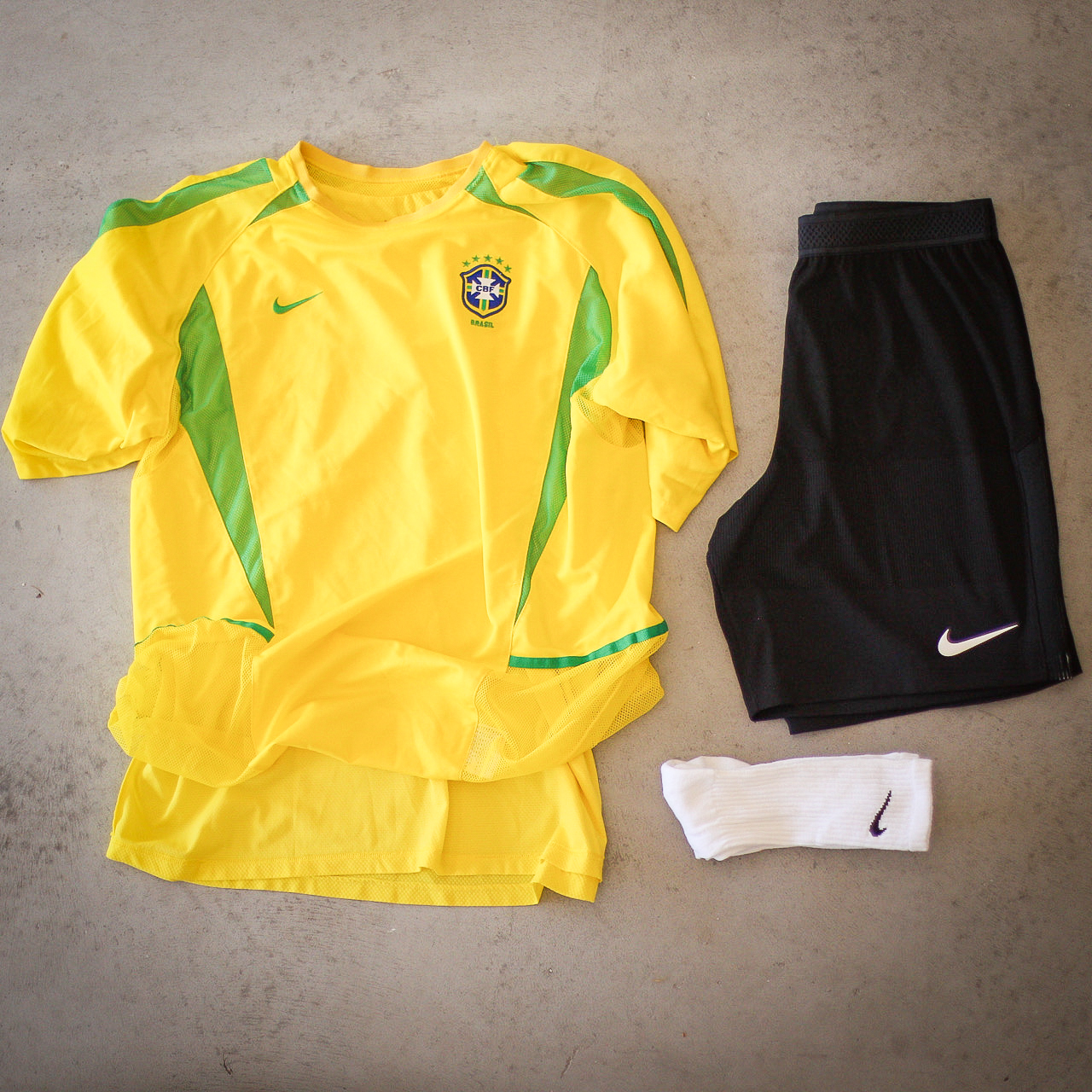 Yellow Brazil football shirt with green detailing, black Nike shorts and white socks laid out on concrete